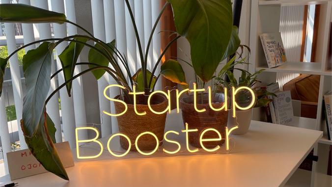 A photo of a neon sign reading 'Startup Booster' in front of a few potted plants in an office setting.